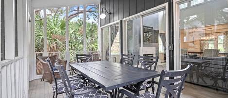 An outdoor patio with a wooden table and chairs, large windows, and a view of greenery.