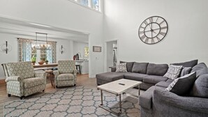 Een ruime woonkamer met een grijze modulaire bank, gestreepe fauteuils, een bijzettafeltje en een grote wandklok.
