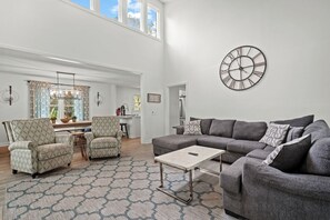Ein geräumiges Wohnzimmer mit einem grauen Ecksofa, gemusterten Sessel, einem Couchtisch und einer großen Wanduhr.