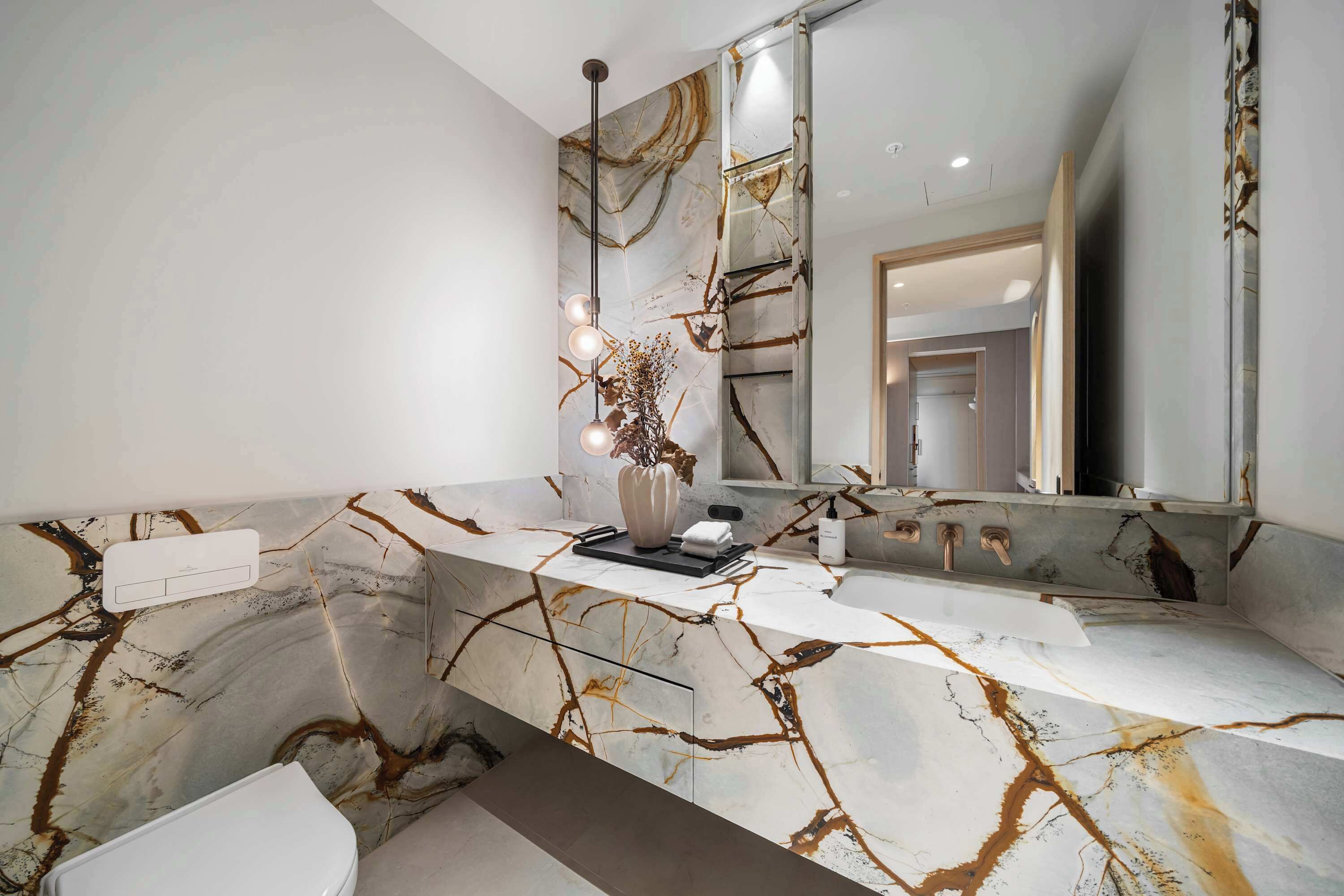 Une salle de bain avec un comptoir en marbre, un grand miroir et un vase contenant des fleurs séchées.