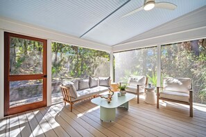 Ein sonnendurchflutetes Zimmer mit Holzboden, einer Couch, einem Tisch und großen Fenstern mit Blick auf einen Wald.