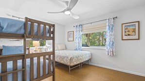 Un dormitorio con una cama litera, una cama individual, un ventilador de techo, una ventana con cortinas y un cuadro en la pared.