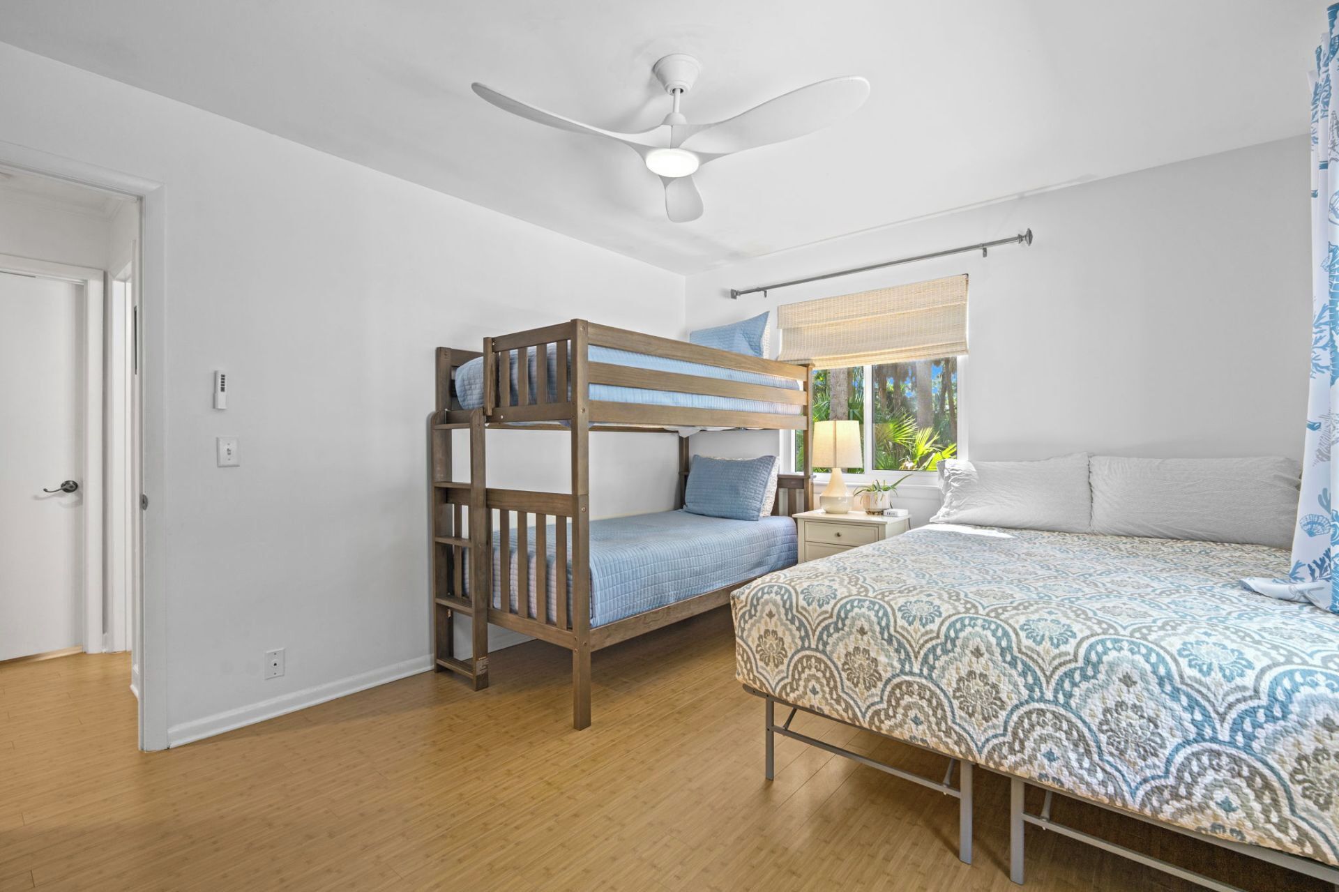 A bedroom with a bunk bed, a single bed, a ceiling fan, a window with a view, and a bedside table with a lamp.