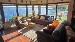 A spacious living room with a large window overlooking a body of water, furnished with a sofa, ottoman, and a hanging chair. - Best Views of Webster Lake! Paddleboards + Swimming (North Webster)