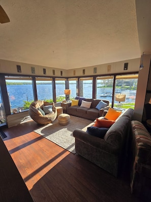 A spacious living room with a large window overlooking a body of water, furnished with a sofa, ottoman, and a hanging chair.