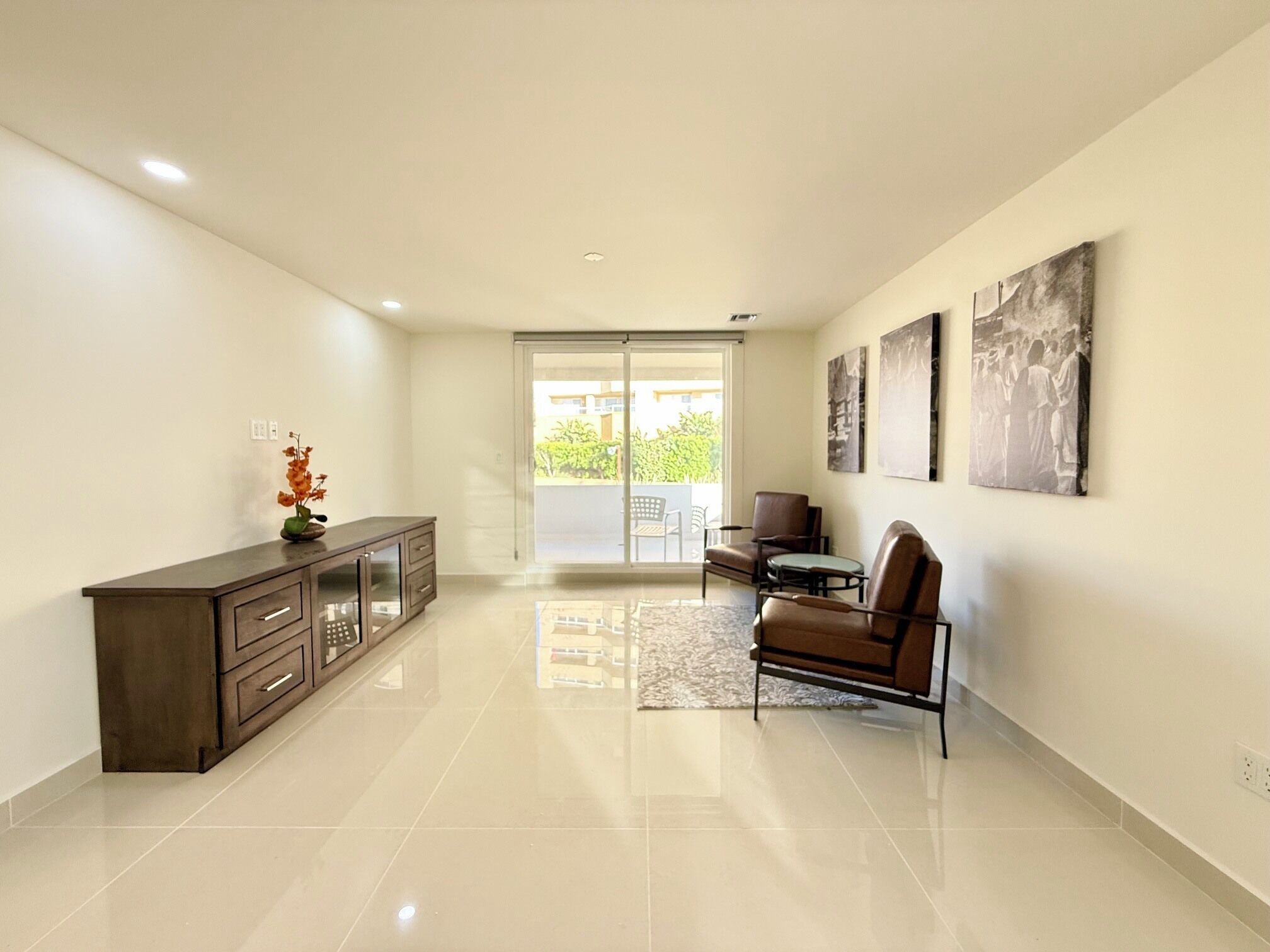 A spacious living area with a sliding glass door leading to a balcony, a dark wooden cabinet, and a brown leather chair.