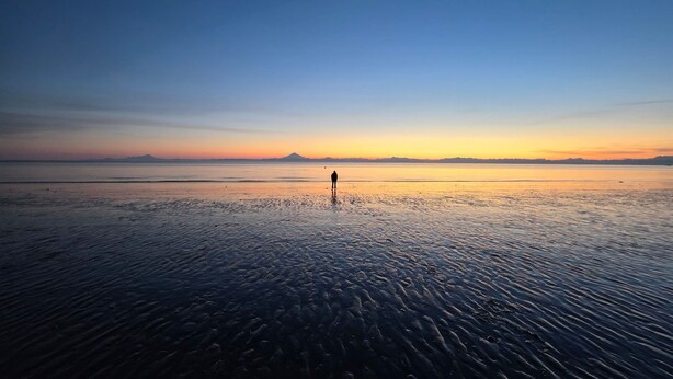Una playa con una persona de pie en el agua, un mar tranquilo y una montaña al fondo.