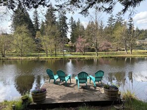 Une terrasse en bois avec quatre chaises Adirondack vertes, au bord d’un lac paisible, entourée d’arbres et avec un belvédère en arrière-plan.