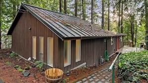 A rustic cabin with a sloped roof, wooden siding, and large windows, surrounded by a forested area.