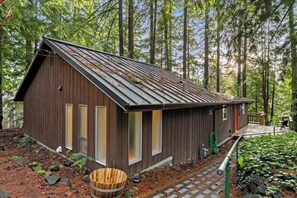 A rustic cabin with a sloped roof, wooden siding, and large windows, surrounded by a forested area.