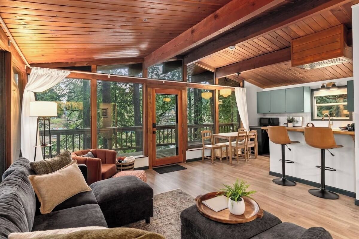 Un vaste espace de vie comprenant un canapé, un pouf et une table à manger avec des chaises. Il y a également un coin cuisine avec des tabourets de bar et une grande fenêtre donnant sur une terrasse.