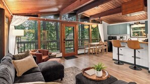 A spacious living area with a sofa, ottoman, and a dining table with chairs. There is a kitchen area with bar stools and a large window overlooking a deck.