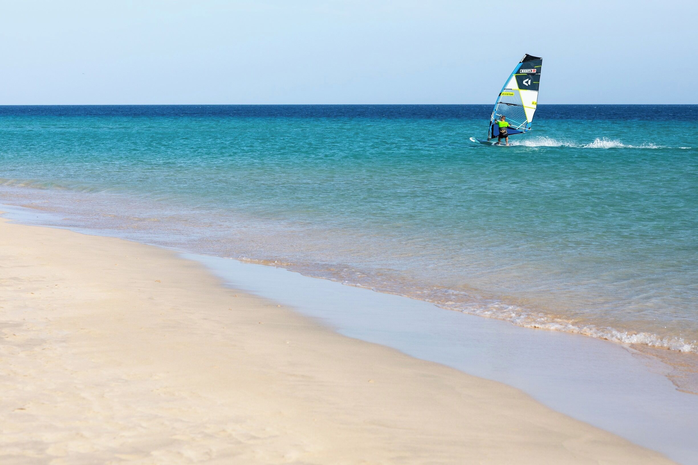 A beach with clear blue water and a person windsurfing.