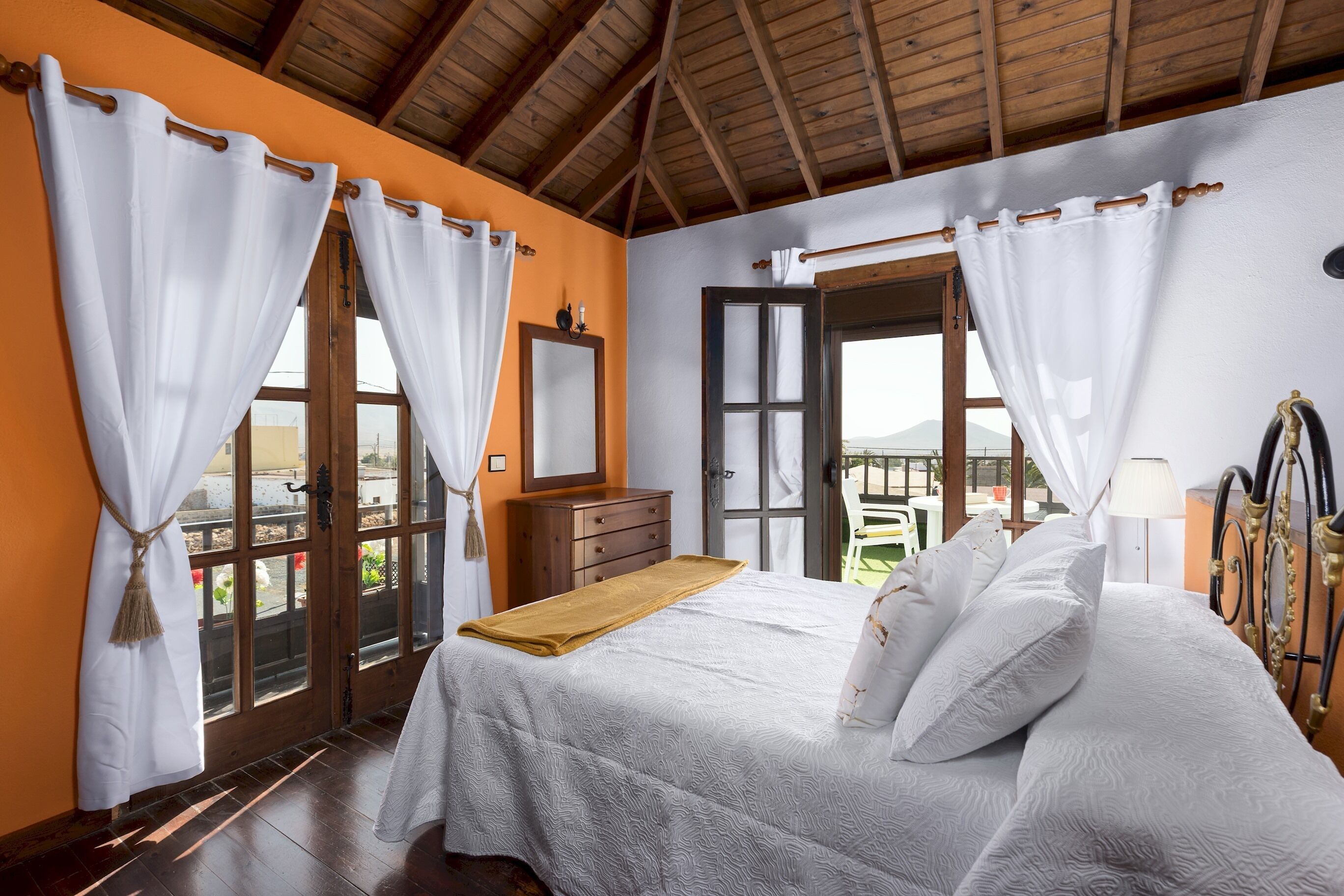 A bedroom with a bed, wooden headboard, white curtains, a wooden dresser, and a view through an open door.