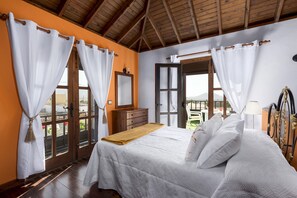 Une chambre à coucher avec un lit, une tête de lit en bois, des rideaux blancs, une commode en bois et une vue sur l’extérieur par une porte ouverte.