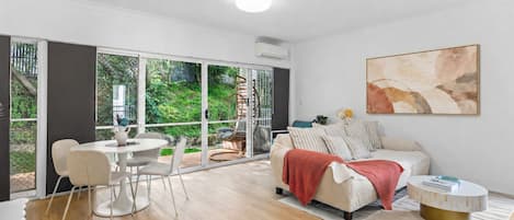 A modern living room with a sofa, a dining table with chairs, a large window, and a painting on the wall.