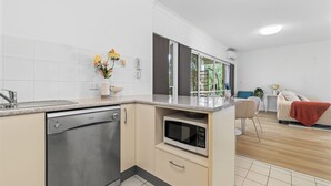 A modern kitchen with a dishwasher, microwave, and a small dining area with a chair and table. - 2BR 2BA Indooroopilly | Walk to Shops & Bus (Indooroopilly)