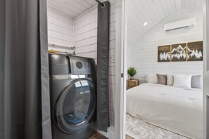 A compact room with a washing machine, a bed with white bedding, and a wall-mounted air conditioner. - Private Tiny Home Surrounded By Farmland W/Hottub (Gordonville)