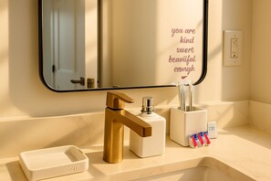 Un baño con grifo de oro, espejo, jabonera y porta cepillos de dientes con pasta dental.