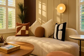 Una sala de estar acogedora con un sofá, cojines, una pequeña mesa redonda con un libro, una planta en maceta y una lámpara de pared.
