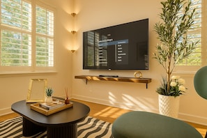 A modern living room with a flat-screen TV mounted on the wall, a round coffee table, a bookshelf, and a potted plant.