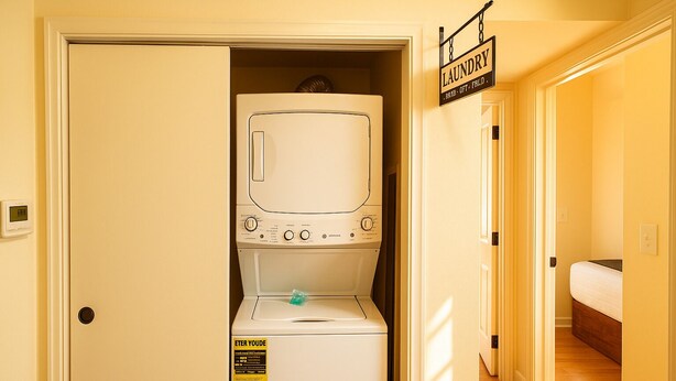 A laundry room with a front-loading washer and dryer, a sign above the dryer, and a bed visible in the adjacent room.