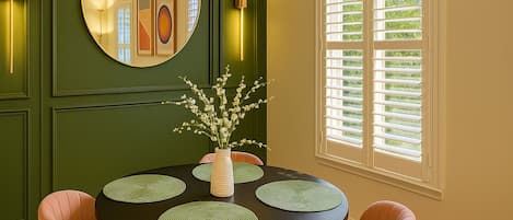 Un comedor con una mesa redonda, sillas, un florero con flores, un espejo y una ventana con persianas.