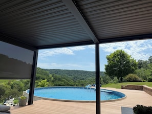 A covered outdoor area with a swimming pool, wooden deck, and a view of a forested hillside.