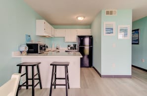 Una cocina con isla central, armarios blancos, un refrigerador negro y dos taburetes de bar.