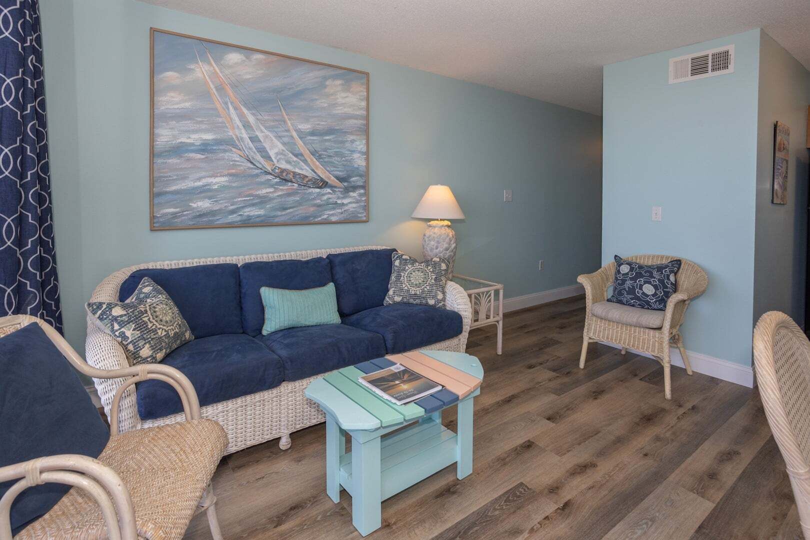 Un salon avec un canapé bleu, des fauteuils en osier, une table basse et un tableau représentant des voiliers.