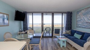Una habitación con una mesa de comedor, sillas, un sofá, un televisor y un ventanal con vistas a la playa.