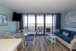 Quarto com mesa de jantar, cadeiras, sofá, televisão e uma grande janela com vista para a praia.