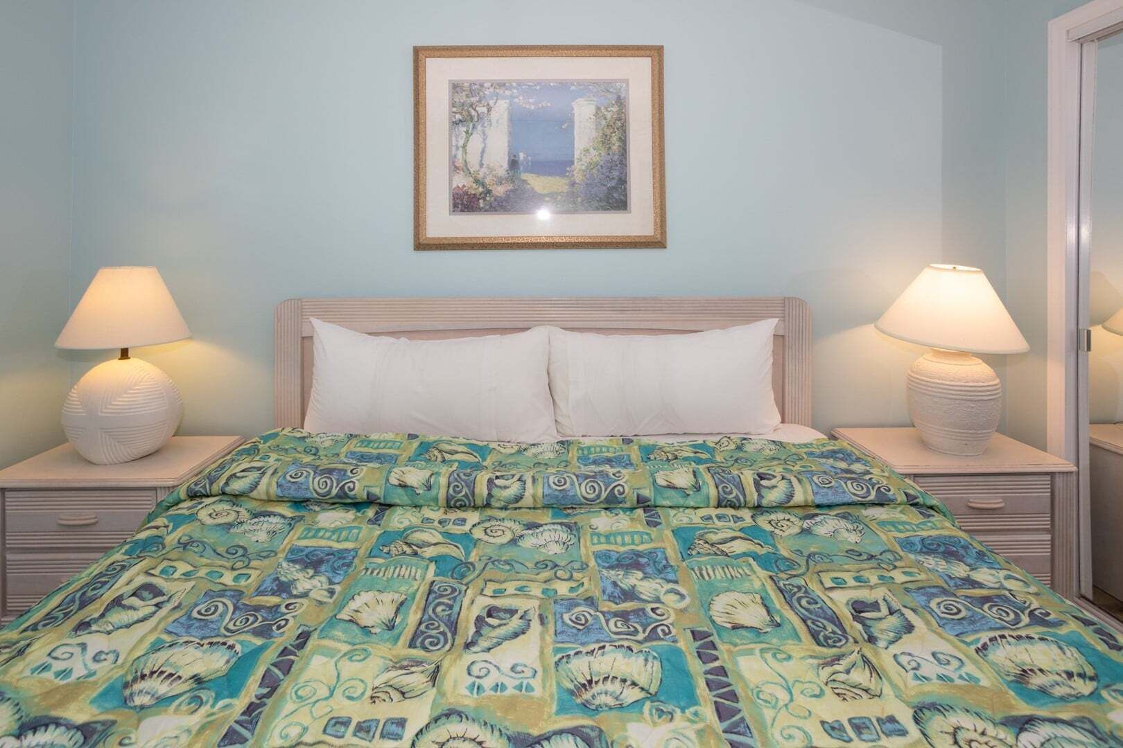 Un dormitorio con una cama, dos lámparas de noche, un cuadro enmarcado en la pared y una colcha estampada.