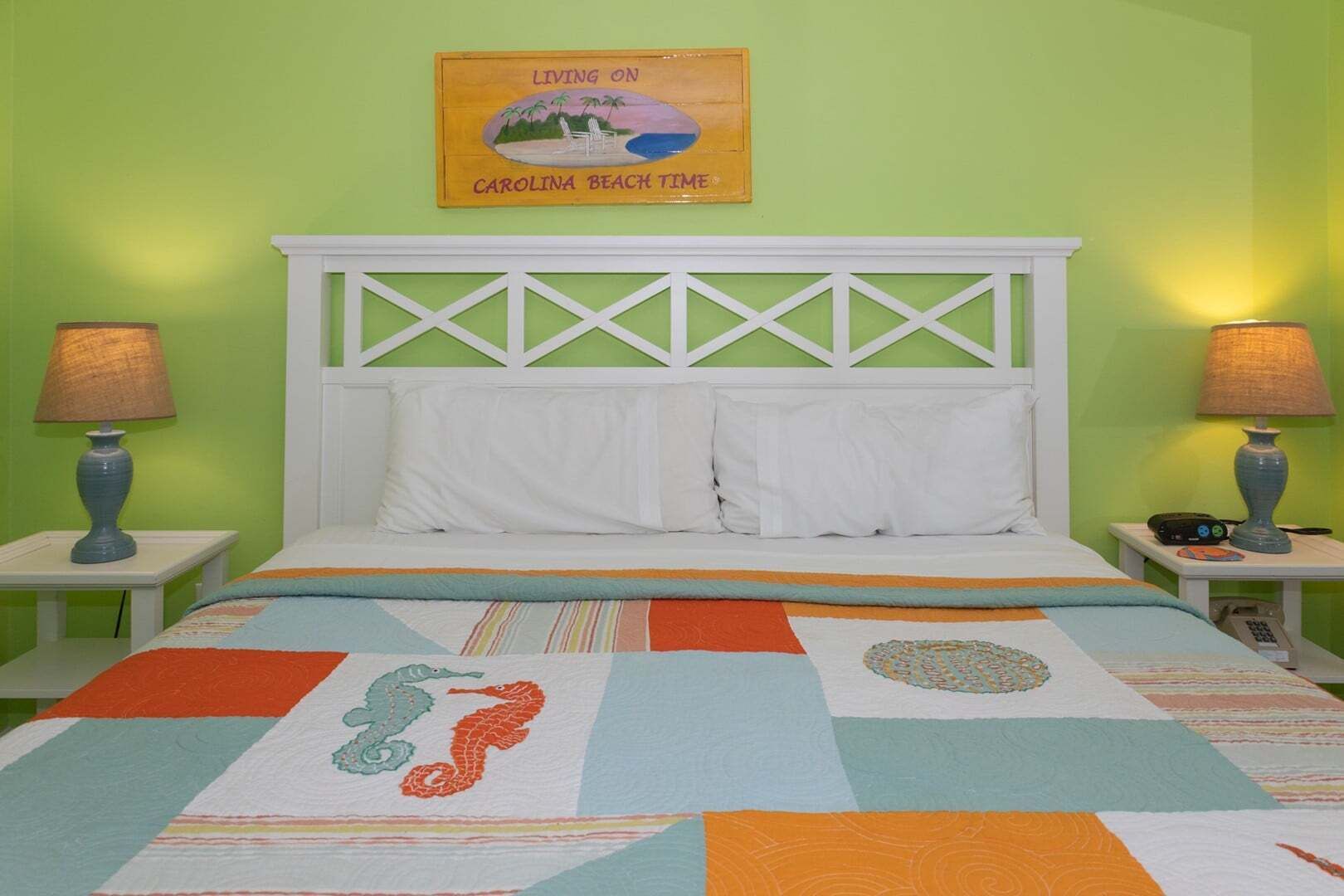 Una cama con cabecera blanca y almohadas, un colcha colorida con diseño de caballito de mar, y un cuadro en la pared.