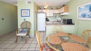 A compact kitchen with a refrigerator, microwave, and a small dining area with wicker chairs and a glass table. - Cozy 1BR oceanfront condo in Carolina Beach 0201 (Carolina Beach)