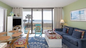 A living room with a sofa, a table, chairs, a TV, and a balcony with a view of the beach. - Cozy 1BR oceanfront condo in Carolina Beach 0201 (Carolina Beach)