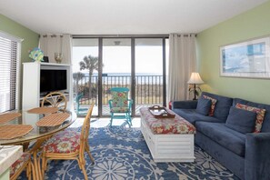 Ein Wohnzimmer mit einer Couch, einem Tisch, Stühlen, einem Fernseher und einem Balkon mit Blick auf den Strand.