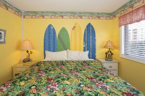 Ein Schlafzimmer mit einem Bett, zwei Nachttischen, einem Fenster mit Jalousien und einem Wandbild im tropischen Stil.