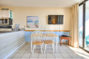 A kitchen with a microwave, stove, and a painting of a beach scene. There is a TV mounted on the wall and a dining area with two chairs and a small table. - Oceanfront escape: 2BR, 2BA condo at Carolina Beach, perfect for a family getaway! (Carolina Beach)