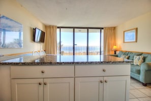 A kitchen with a large island, a dining area with chairs, a sofa, a TV mounted on the wall, and a view of the beach through the sliding glass door. - Oceanfront escape: 2BR, 2BA condo at Carolina Beach, perfect for a family getaway! (Carolina Beach)