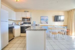 A compact kitchen with a microwave, stove, and dishwasher, a dining area with chairs and a table, and a flat-screen TV mounted on the wall. - Oceanfront escape: 2BR, 2BA condo at Carolina Beach, perfect for a family getaway! (Carolina Beach)