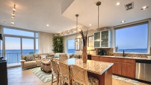 A spacious living area with a large window overlooking the sea, a dining area with a kitchen in the background, and a cozy seating area with a view.