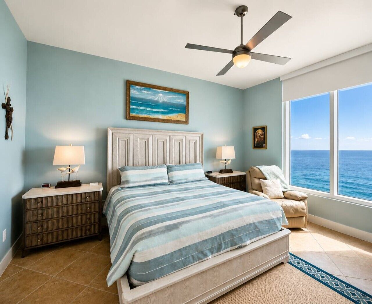 Une chambre à coucher avec un lit, des tables de chevet, un ventilateur de plafond, une fenêtre donnant sur l’océan et un tableau accroché au mur.