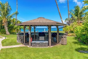 Ein Kiosk mit Steinunterbau und Metallveranda, der einen Blick auf einen Strand mit Palmen und einem wolkenlosen, blauen Himmel bietet.