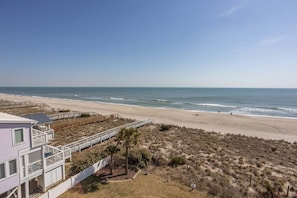 Blick auf einen Strand mit einem Haus, einer Holzterrasse und feinem Sand.