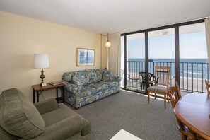 Ein Zimmer mit einer Couch, einem Sessel, einem Tisch, einer Lampe, einem Bilderrahmen und einem Balkon mit Meerblick.