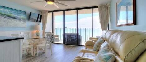 A room with a ceiling fan, a beige sofa, a dining table with chairs, and a view of the ocean through large windows.
