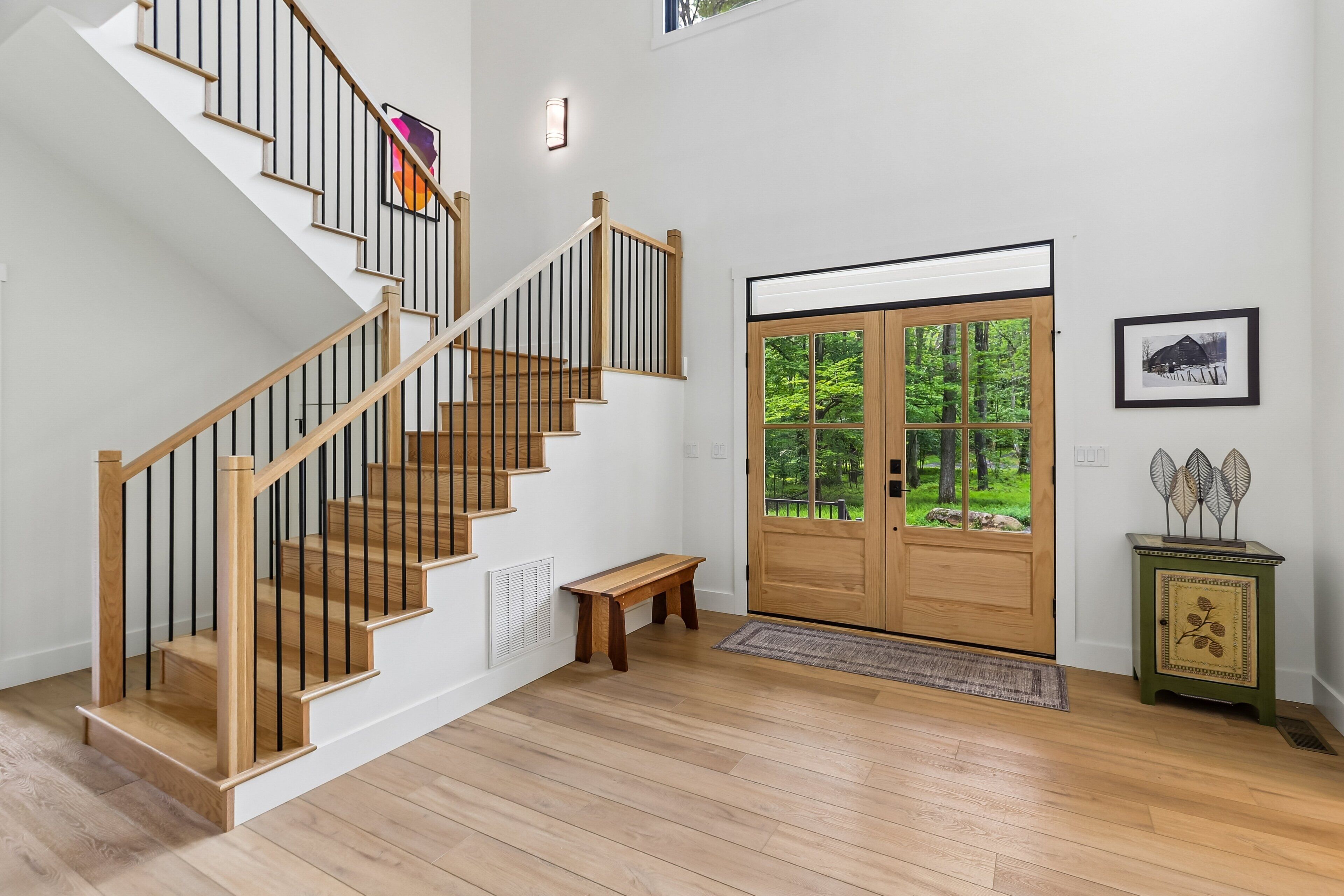 A staircase with wooden railings and a bench, a wooden door with a view of greenery, and a small cabinet with decorative items.
