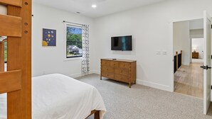 Un dormitorio con cama, cómoda, televisor y ventana con vista a una casa.