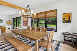 A dining area with a large wooden table, multiple chairs, a hanging light fixture, and a view of the outdoors through large windows. - Experience luxury at Summit and Shore, a 5-bedroom retreat in beautiful Swanton, MD. (Swanton)